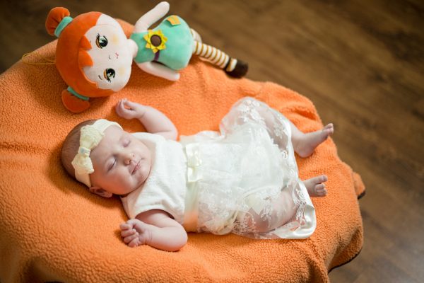 baby in a white dress and bow laying on an orange blanket next to a doll