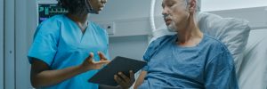 older man lying in a hospital bed listening to his nurse explain something using an ipad