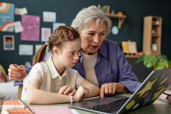 Senior woman guiding young girl with Down syndrome on laptop while sitting at a table in cozy home office, surrounded by educational materials